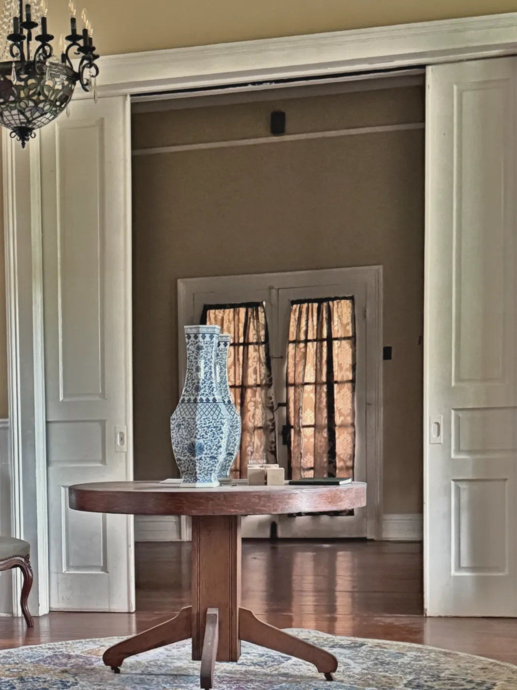 Tall paneled pocket doors open to a parlor with a pedestal table and curtained French doors